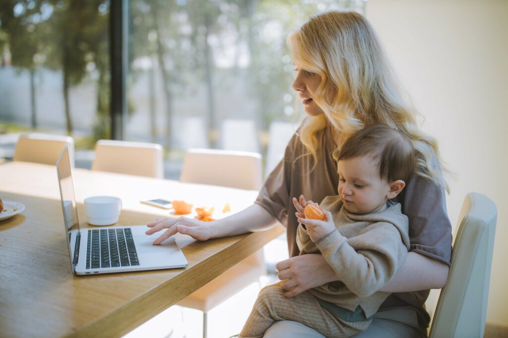 Woman carries her baby while she works at home on her laptop.