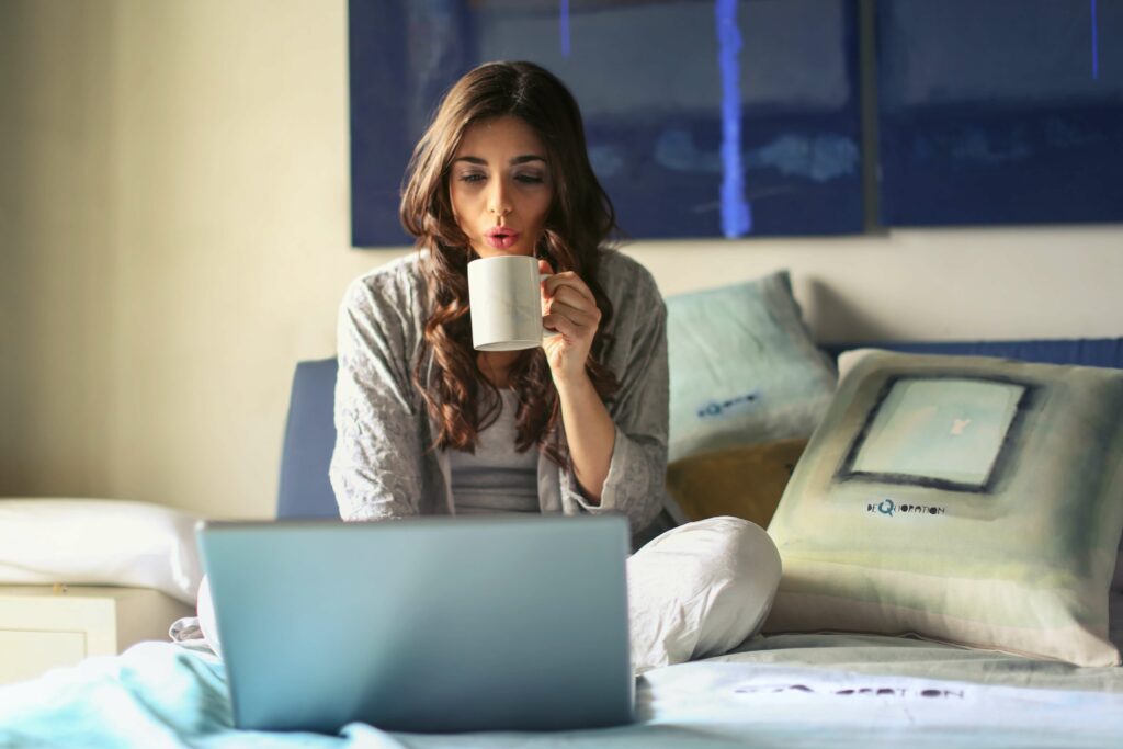 Work from home woman sips coffe on her bed as she looks at her laptop.