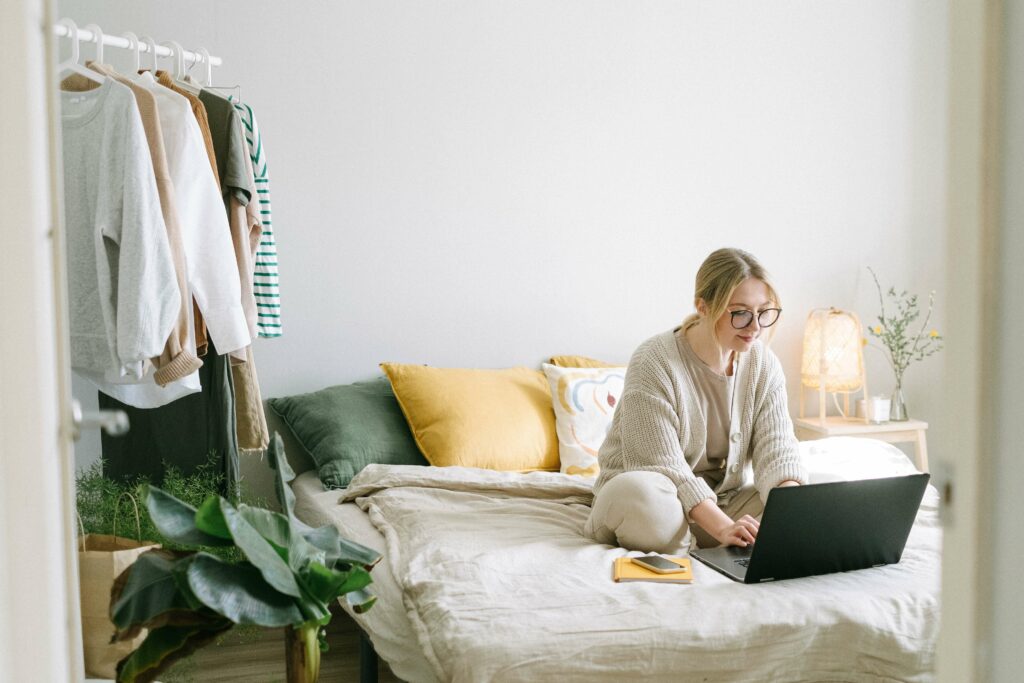 Woman works from home while sitting in bed with a clothes rack on the background.