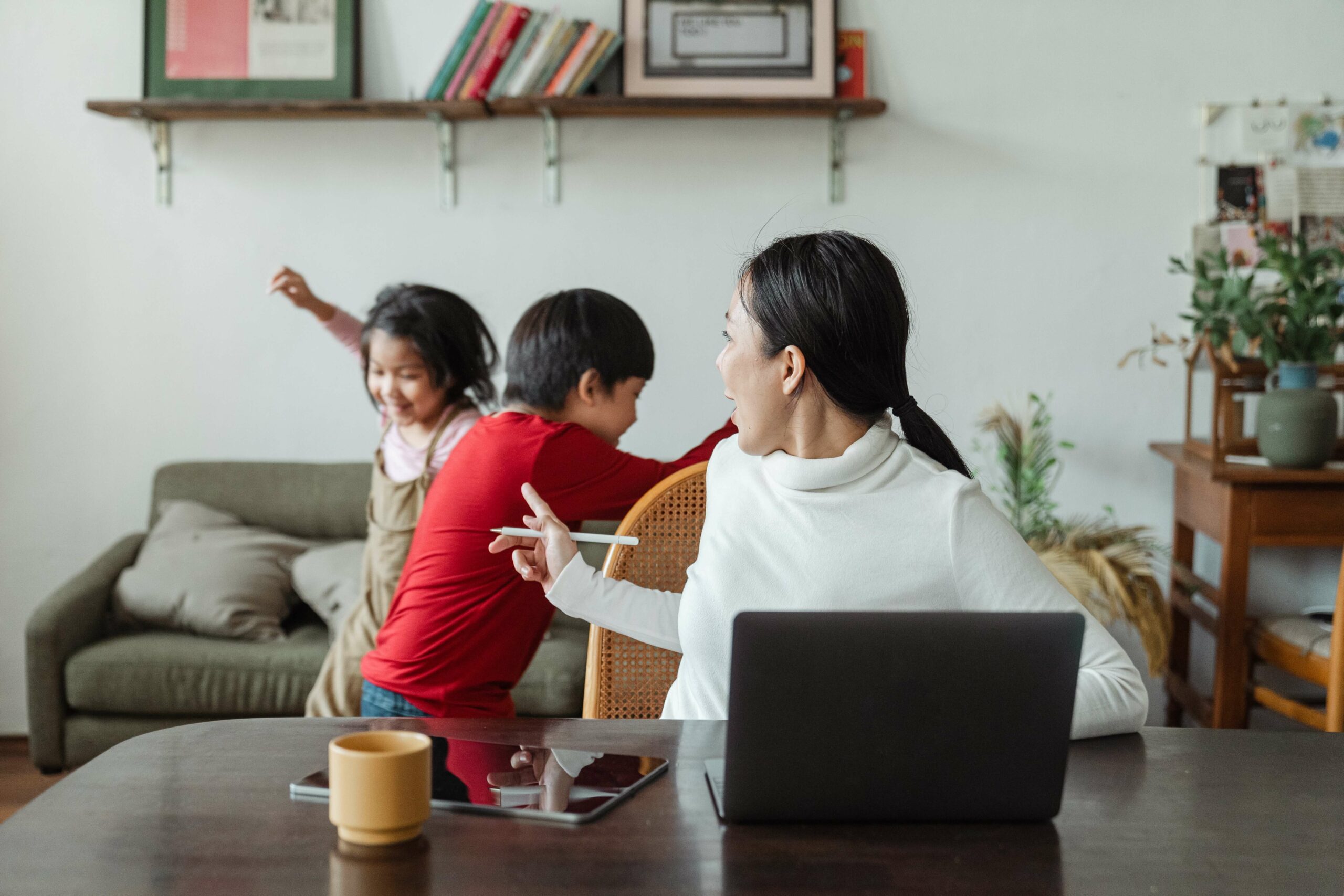 Mother who is work at home talks to her two kids playing behind her as she sits in front of her laptop.