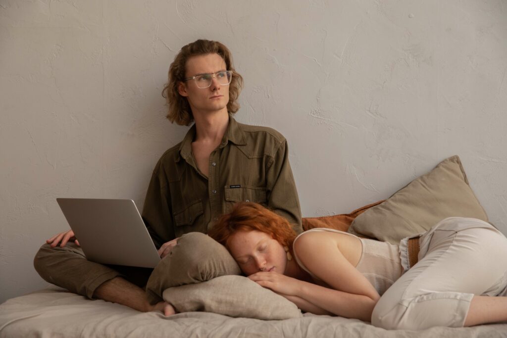 Remote working man works on his laptop in bed while a woman sleeps on his thigh.